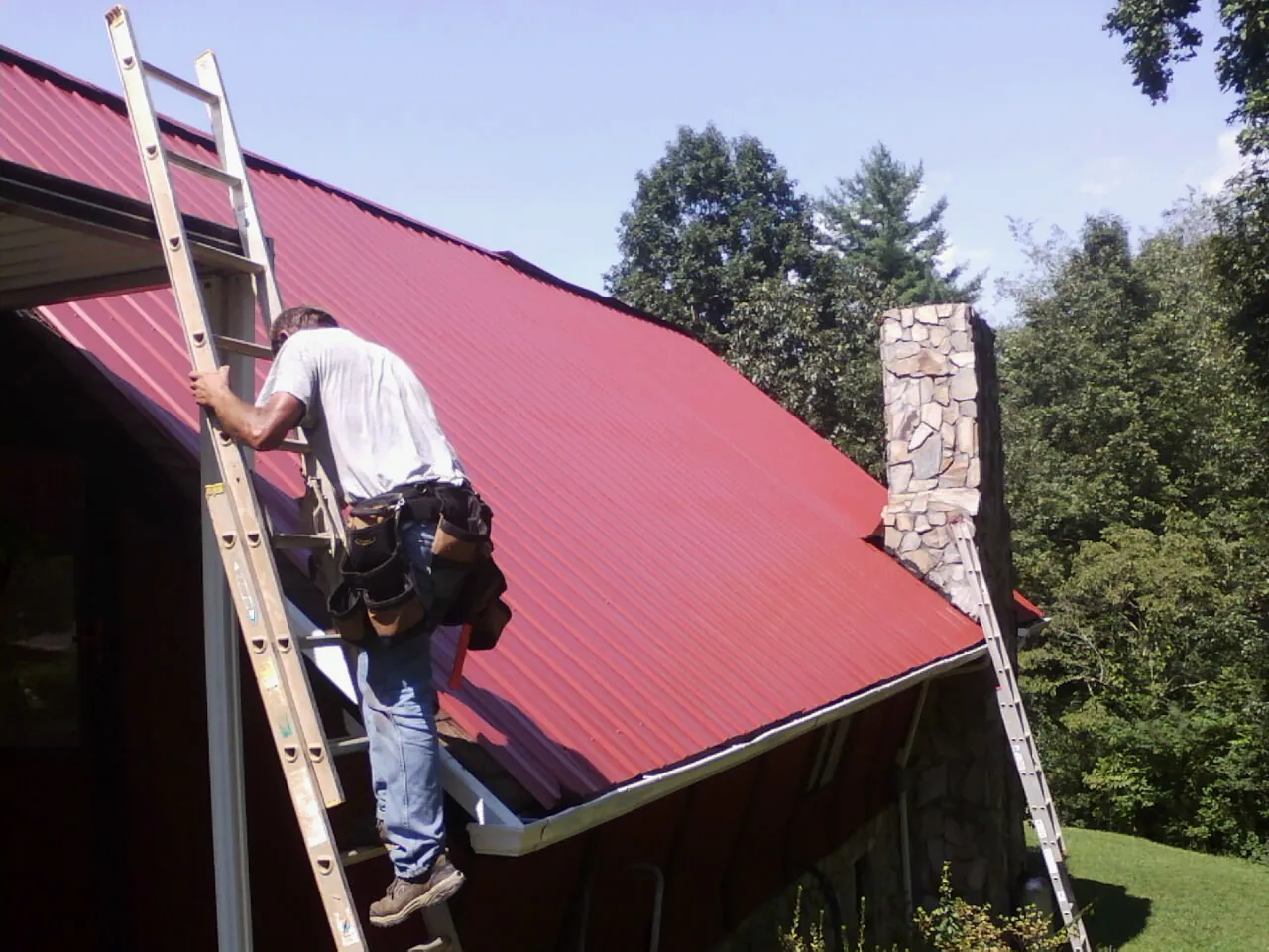 Roofer on a ladder beside a red metal roof and stone chimney — Soffit & Fascia Repair by Roofing Brothers in Greenbush